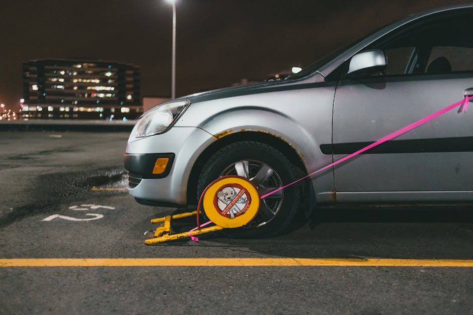 A silver hatchback car is parked in a multi-storey car park at night, with a yellow wheel clamp securing the front right wheel to prevent removal. The clamp features a circular sign with a cartoon dog
