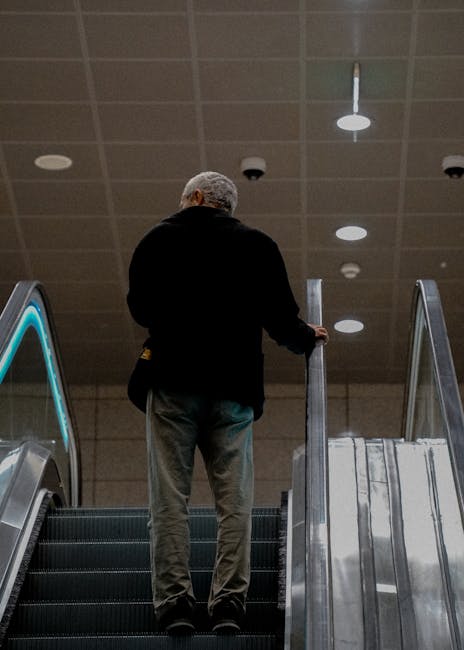 A downward view of an escalator in an indoor setting, with metallic steps and black rubber handrails on either side. The surrounding walls are smooth and glossy with a reflective surface, lined with w