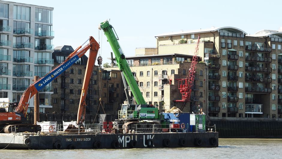 A construction barge positioned on a river features two large cranes, one orange and one green, actively engaged in lifting and moving equipment or materials. The orange crane, marked with 'LAND & WAT