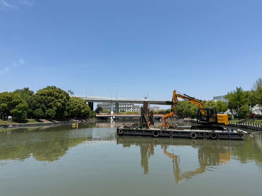 A construction barge positioned on a river features two large cranes, one orange and one green, actively engaged in lifting and moving equipment or materials. The orange crane, marked with 'LAND & WAT
