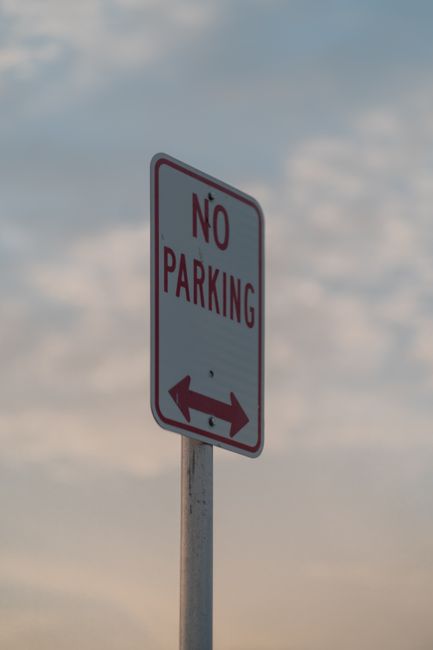 A vertical metal pole supporting a rectangular white 'No Parking' sign with red text and border, displaying a double-ended arrow at the bottom indicating no parking in either direction. The sign is po